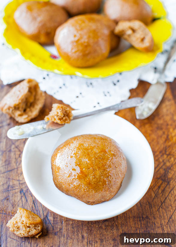 A no-knead whole wheat dinner roll on a white plate next to a platter of rolls. 