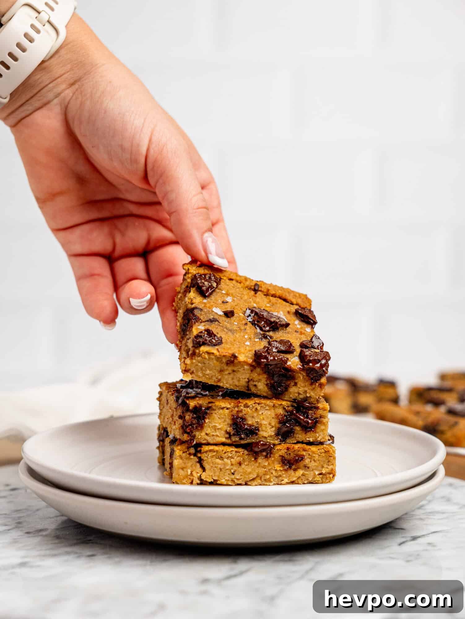 Chewy Chickpea Cookie Bars with Gooey Chocolate Swirls 8 Three chickpea cookie bars stacked on top of each other on a white plate, a hand is picking up the one on top.
