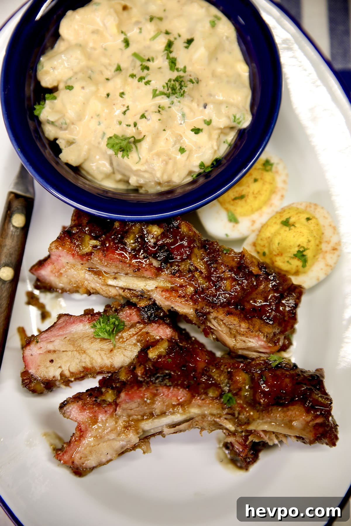 3 ribs on a plate with bowl of potato salad and deviled eggs.