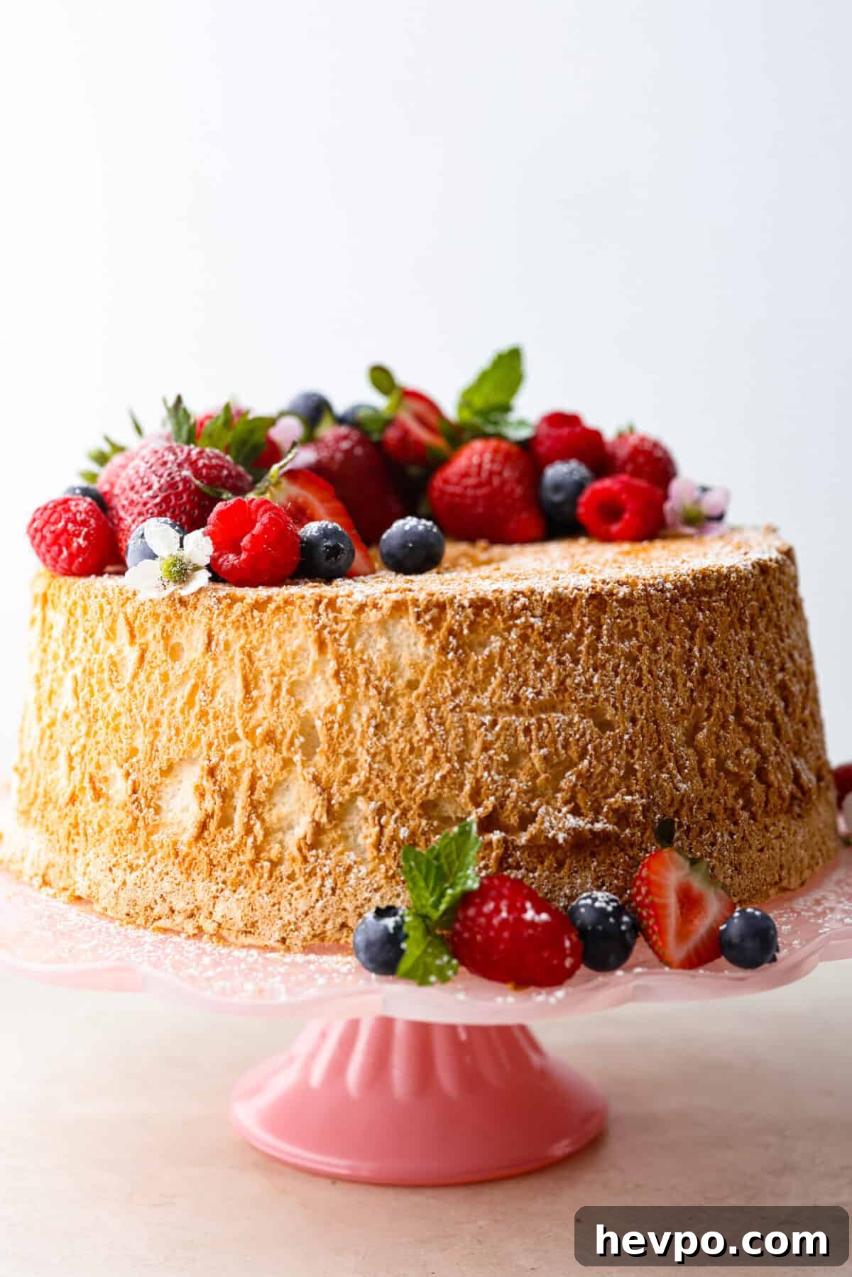 This light & fluffy Angel Food Cake is like a slice of heaven on a plate! It's delicious all on its own but absolutely divine topped with sweetened whipped cream and fresh berries or lemon curd! Side shot of whole angel food cake with berries and blossoms dusted with powdered sugar.