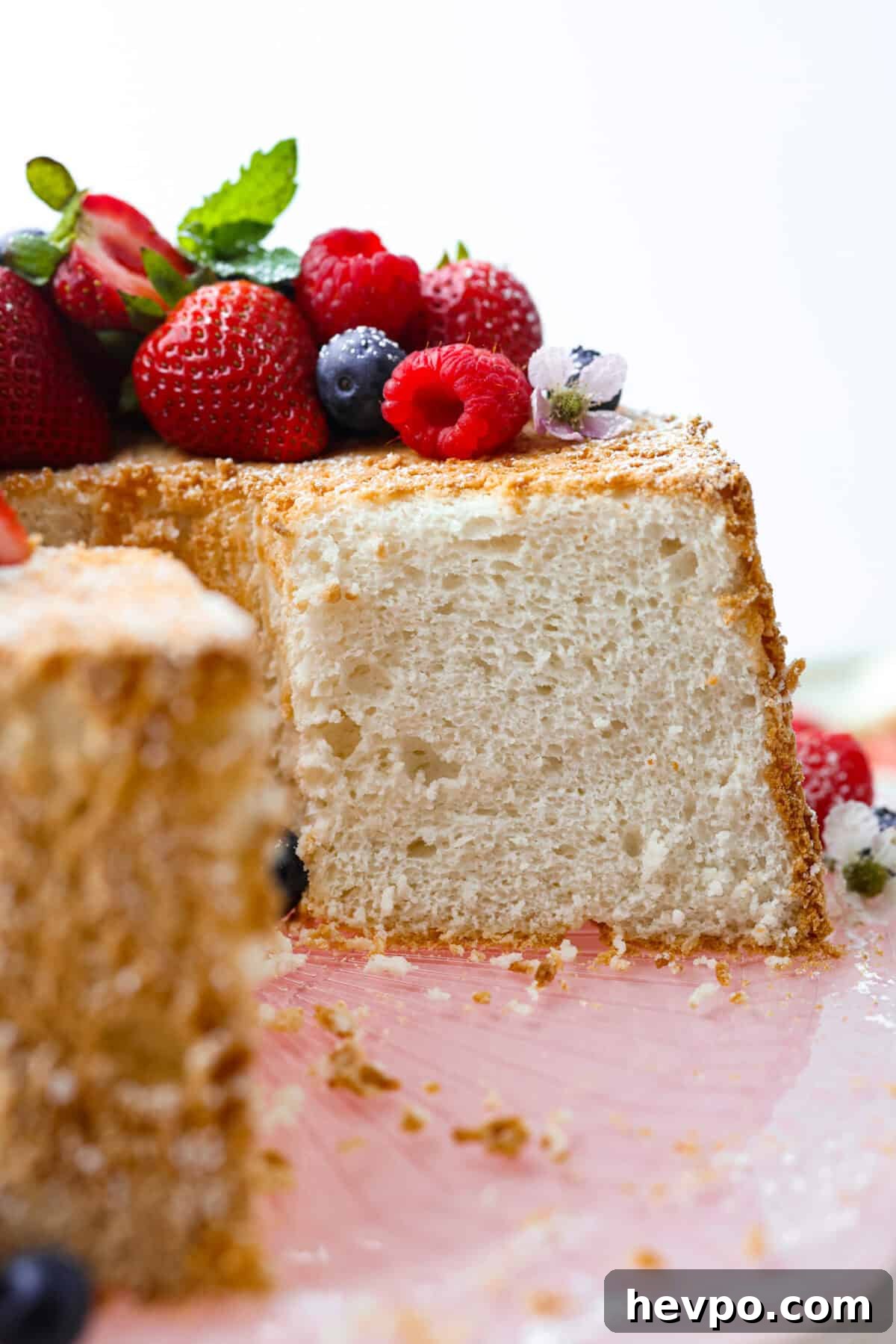 This light & fluffy Angel Food Cake is like a slice of heaven on a plate! It's delicious all on its own but absolutely divine topped with sweetened whipped cream and fresh berries or lemon curd! Side shot of angel food cake covered in berries with a slice taken out of it.