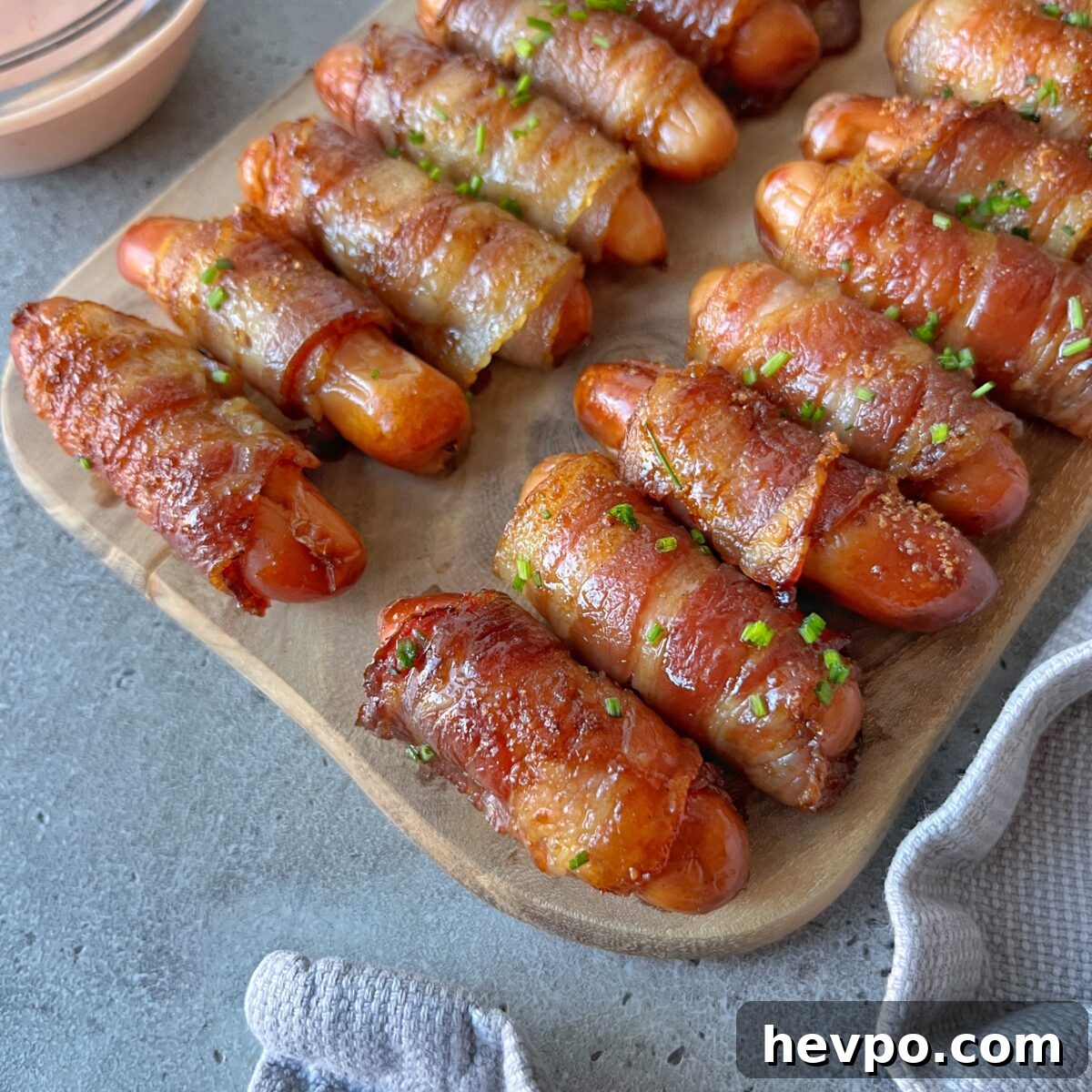 Crispy Bacon-Wrapped Sausages: Crowd-Pleasing Party Snack 2 Mini sausages wrapped in crispy bacon are arranged on a wooden board, garnished with chopped herbs, next to a dipping sauce.