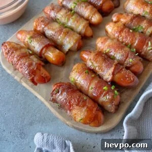 Crispy Bacon-Wrapped Sausages: Crowd-Pleasing Party Snack 10 Mini sausages wrapped in crispy bacon are arranged on a wooden board, garnished with chopped herbs, next to a dipping sauce.