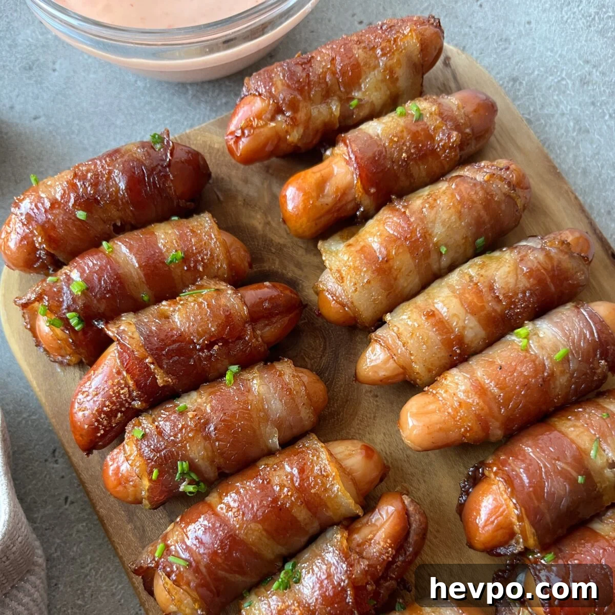 Crispy Bacon-Wrapped Sausages: Crowd-Pleasing Party Snack 3 A wooden board with several bacon-wrapped sausages, garnished with chopped herbs, next to a bowl of dipping sauce.