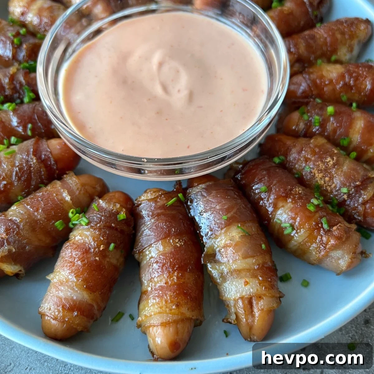 Crispy Bacon-Wrapped Sausages: Crowd-Pleasing Party Snack 4 A plate of bacon-wrapped mini sausages arranged in a circle around a bowl of creamy pink dipping sauce, garnished with chopped chives.