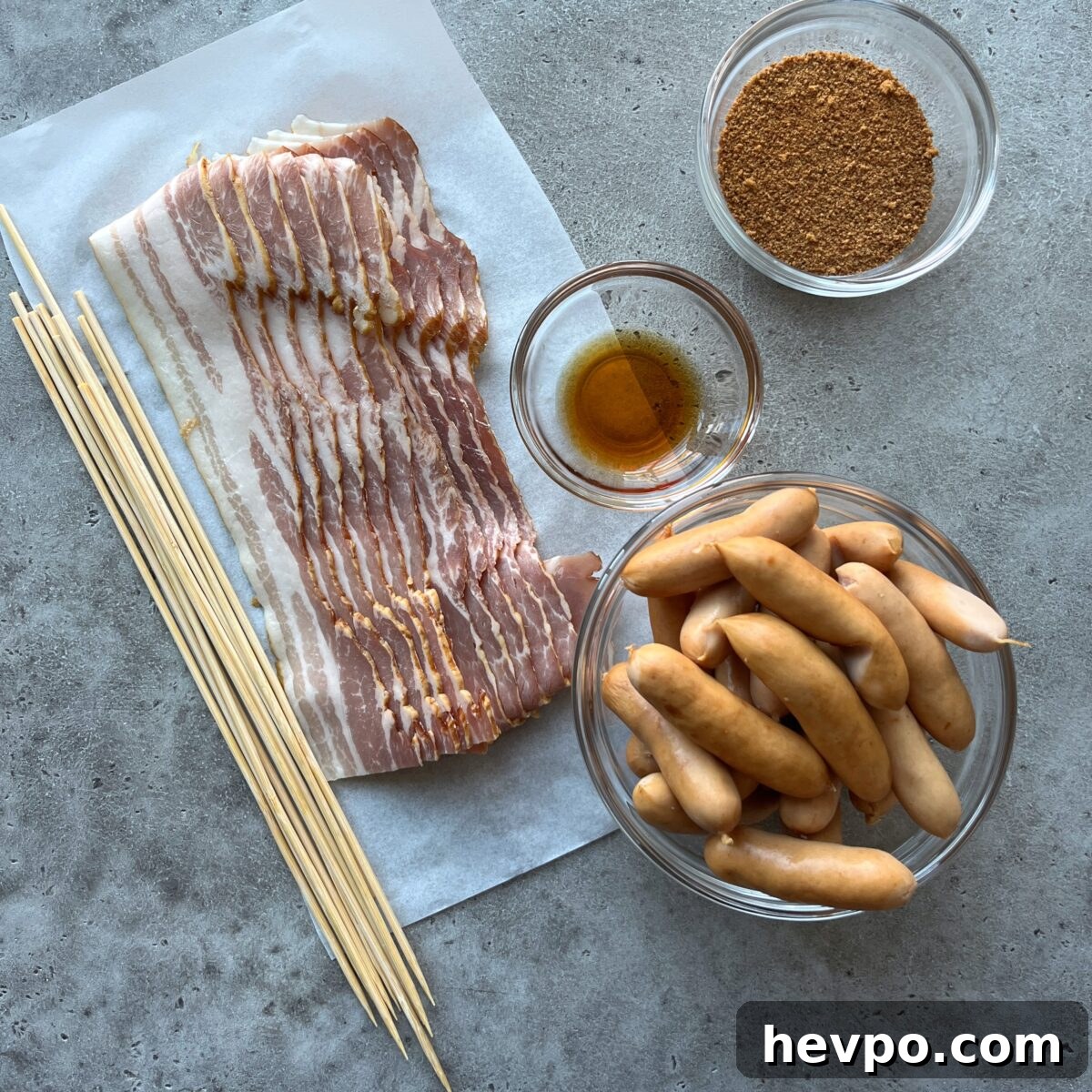 Crispy Bacon-Wrapped Sausages: Crowd-Pleasing Party Snack 5 Sliced bacon, small sausages, wooden skewers, coconut sugar, and a small bowl of maple syrup arranged on a gray surface.