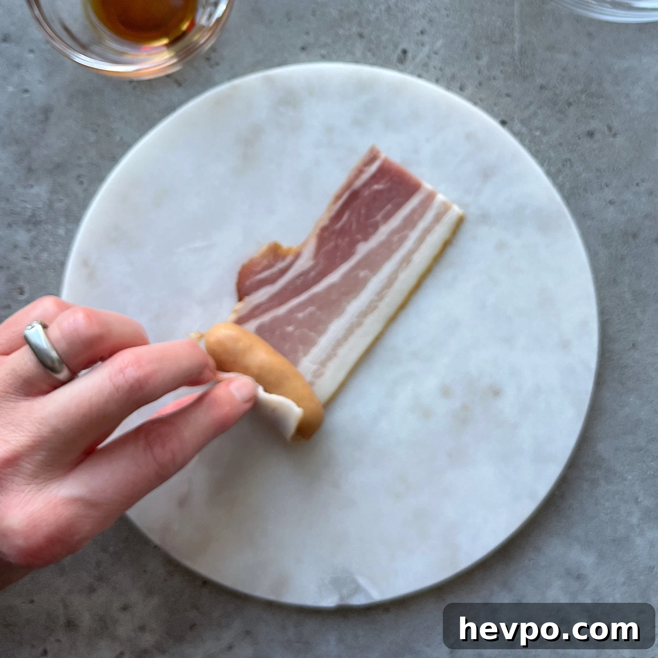 Crispy Bacon-Wrapped Sausages: Crowd-Pleasing Party Snack 6 A hand rolls a sausage in a slice of raw bacon on a round white cutting board, with a small bowl of liquid nearby.