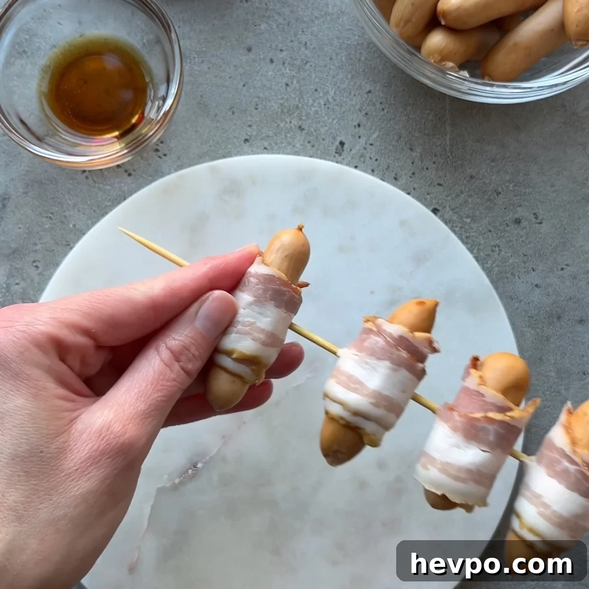 Crispy Bacon-Wrapped Sausages: Crowd-Pleasing Party Snack 7 A hand holds a skewer with small sausages wrapped in bacon, positioned over a round white plate on a gray surface. A bowl of maple syrup and a bowl of sausages are nearby.
