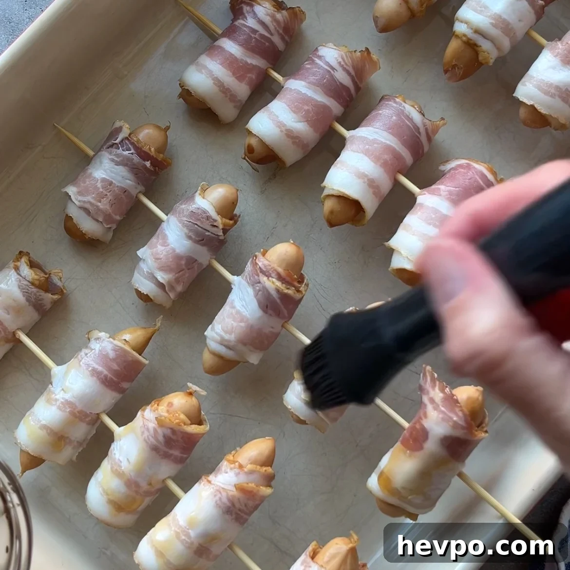 Crispy Bacon-Wrapped Sausages: Crowd-Pleasing Party Snack 8 A hand brushes a maple glaze onto bacon-wrapped sausages arranged on skewers in a baking dish.