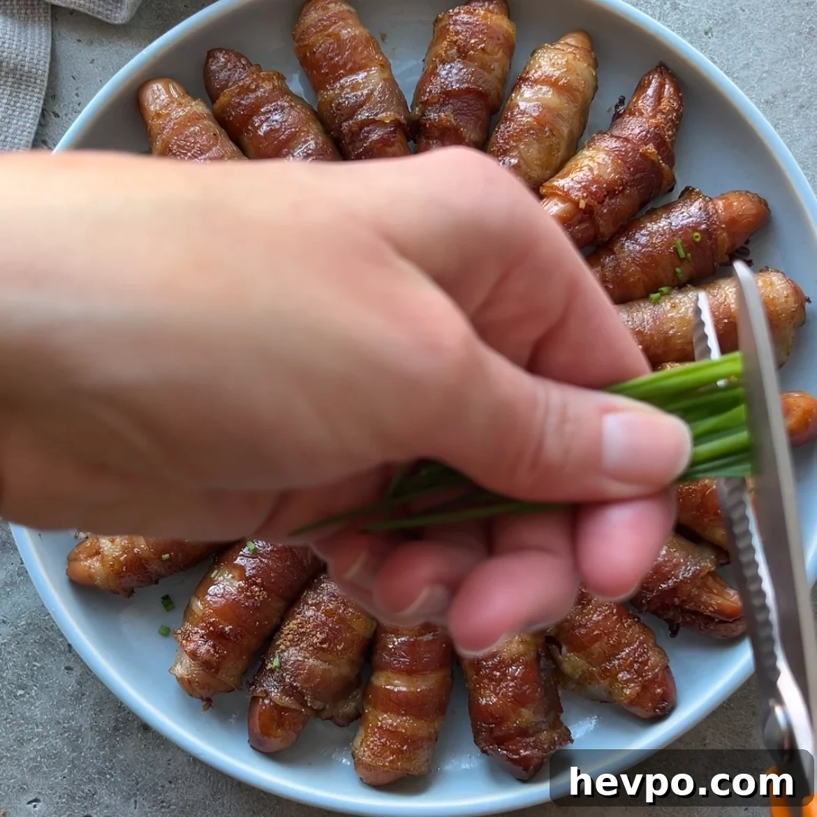 Crispy Bacon-Wrapped Sausages: Crowd-Pleasing Party Snack 9 A hand uses scissors to cut chives over a plate of bacon-wrapped snacks arranged in a circular pattern.