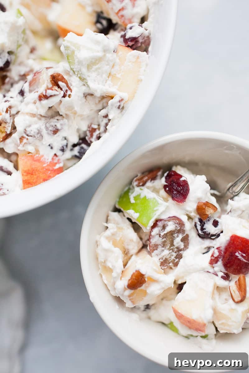 White bowls with creamy whipped cream topped fruit salad with red and green apples, grapes, bananas, cranberries and pecans.