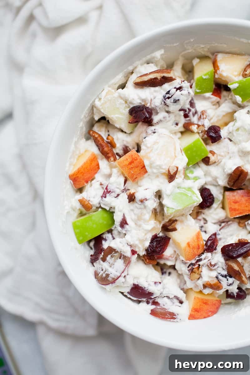 White bowl with creamy whipped cream topped fruit salad with red and green apples, grapes, bananas, cranberries and pecans.