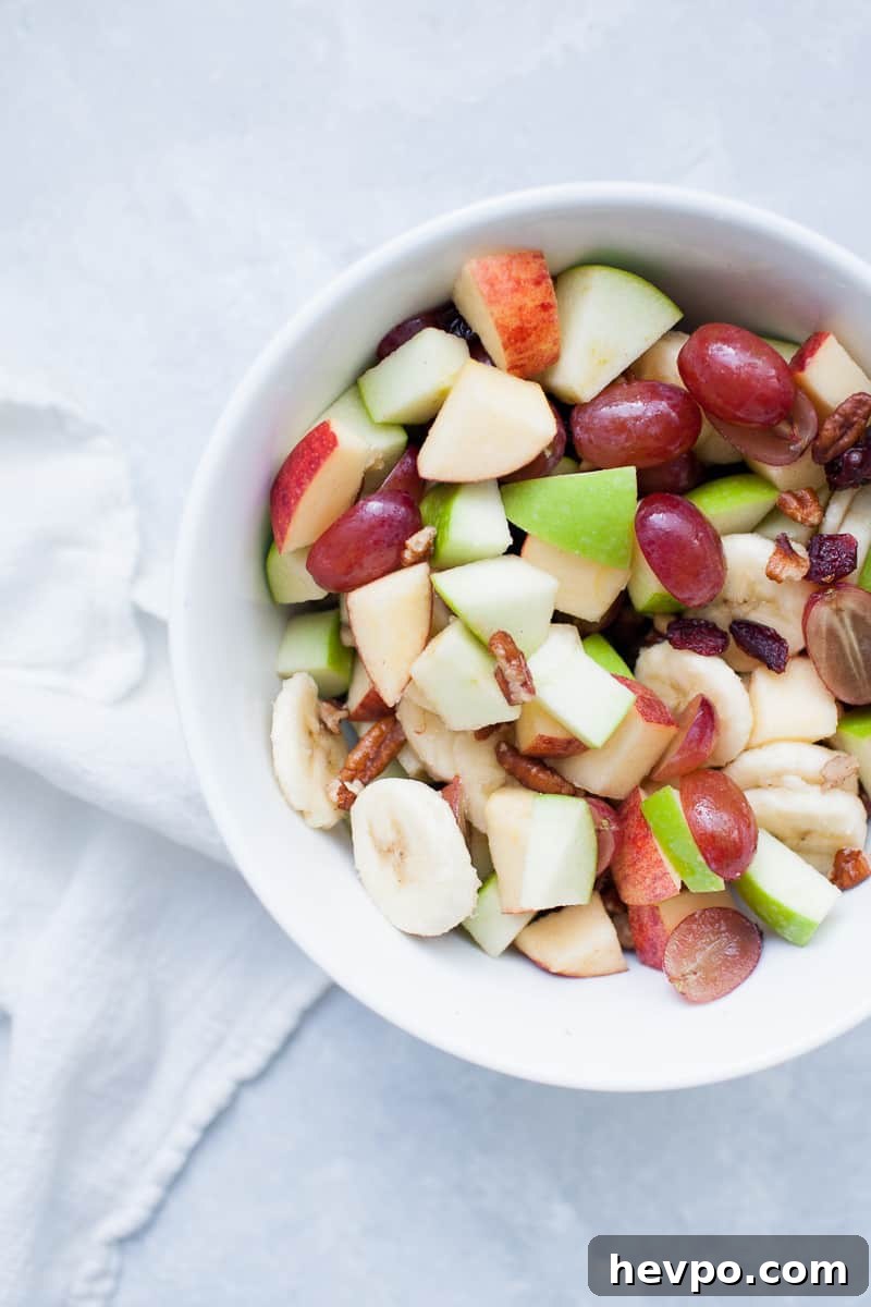 White bowl with red and green apples, pecans, grapes, bananas, and cranberries.