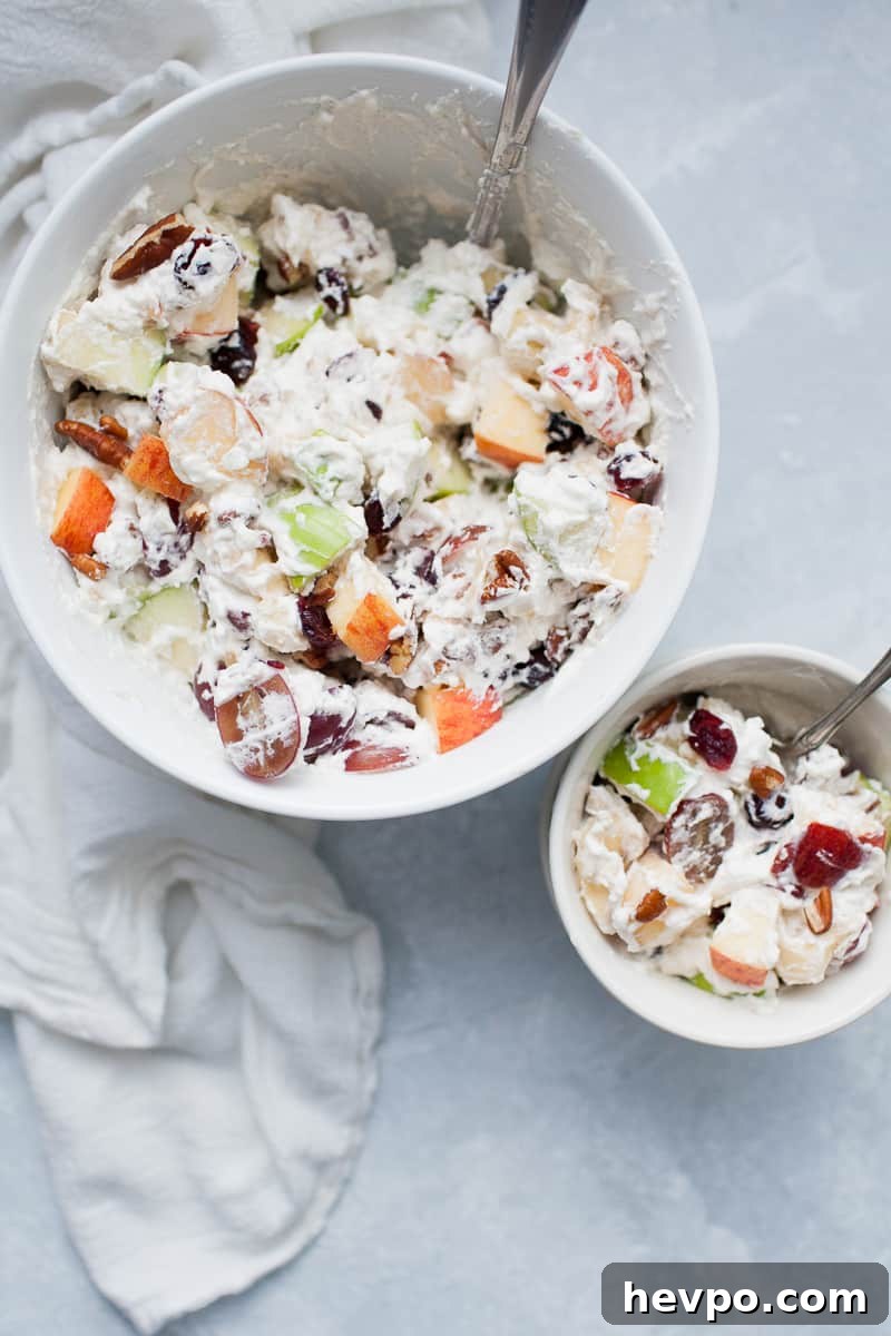 White bowls with creamy whipped cream topped fruit salad with red and green apples, grapes, bananas, cranberries and pecans.