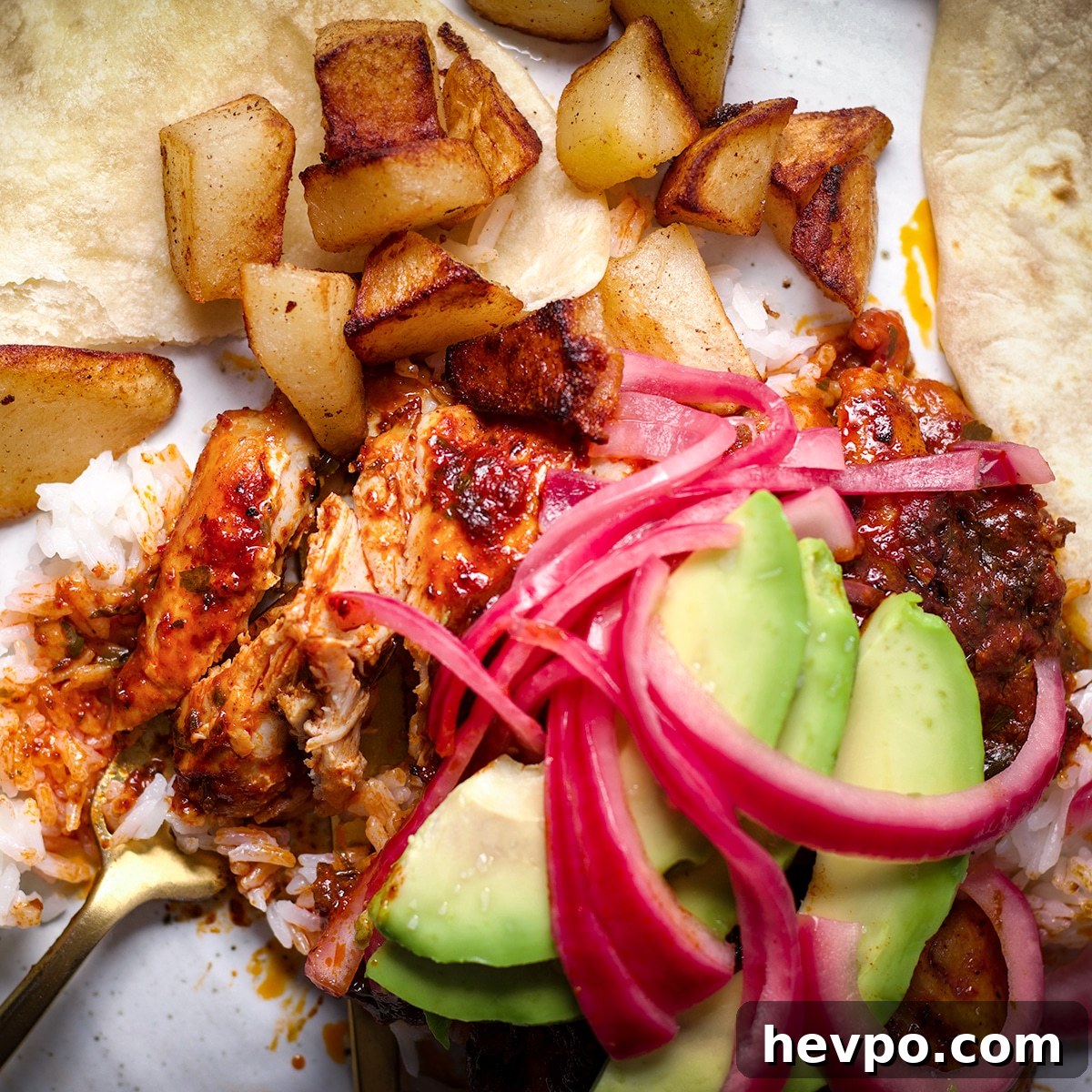 A photo looking down onto a plate of Mexican Adobo Chicken served with rice, crispy fried potatoes and a flour tortilla. A fork has cut into the chicken so you can see the tender, juicy inside of the meat.