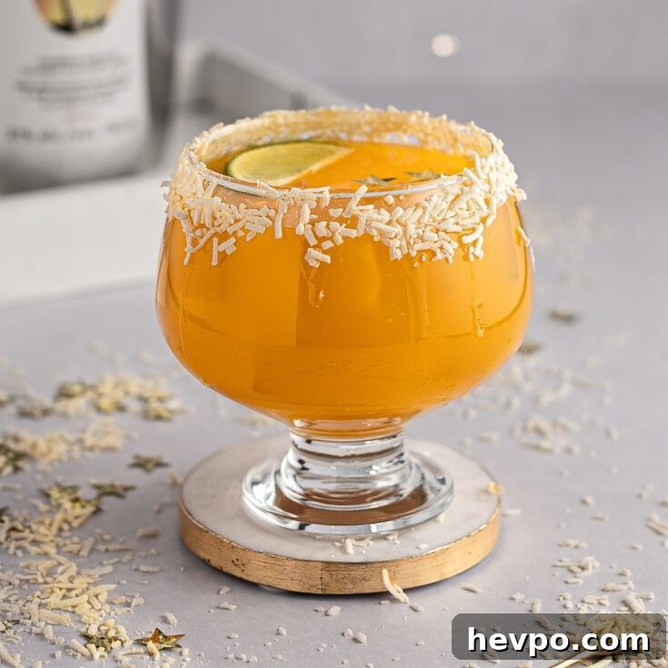 Mango tango drink on with a coconut glass rim, on a white and gold coaster, with gold stars and coconut scattered on the table.