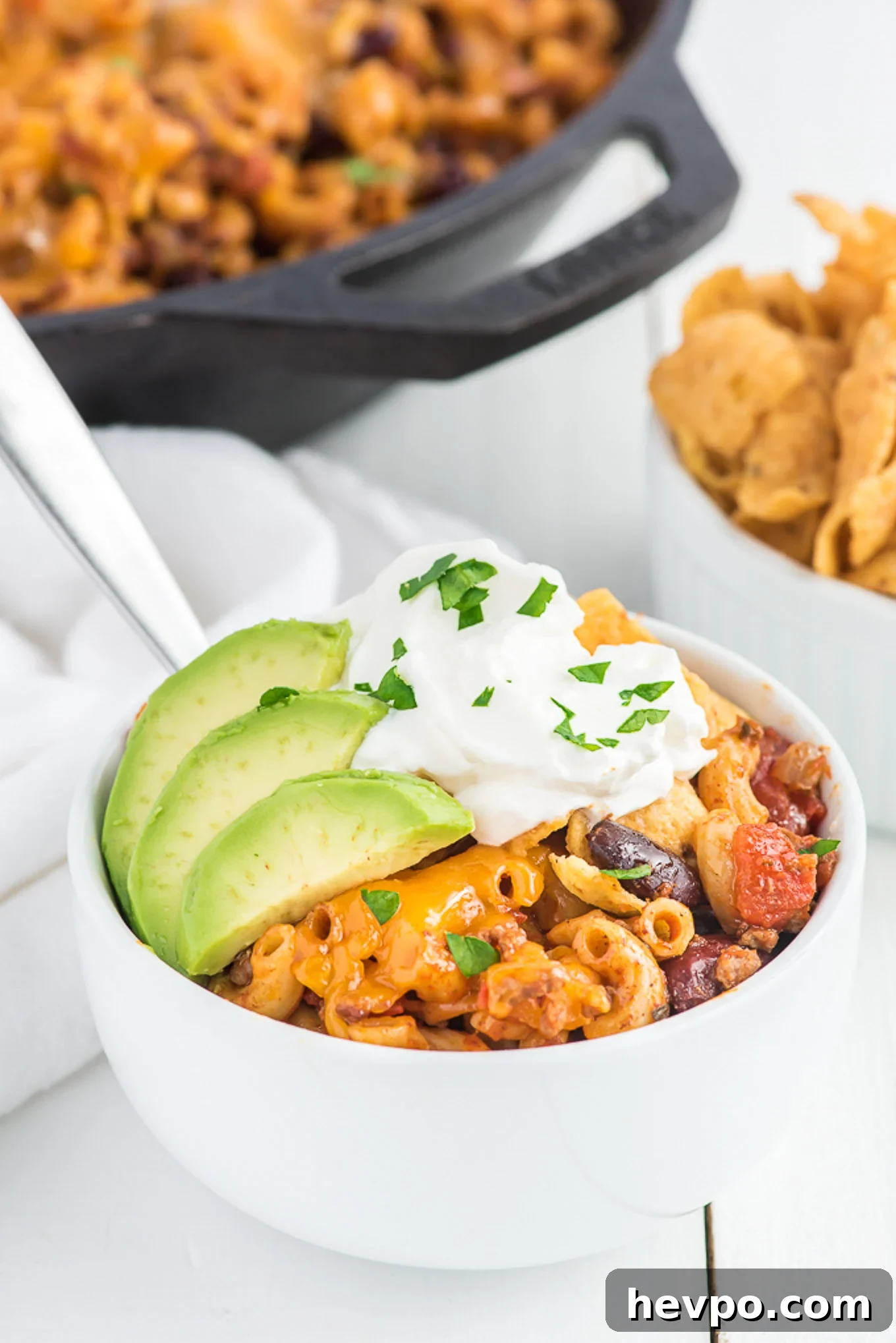 Hearty Chili Mac Recipe: Quick Comfort Food for Weeknights 9 A bowl of chili mac on the table topped with sour cream and avocado with the skillet in the background.
