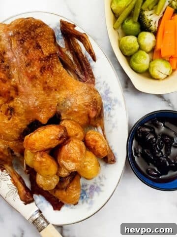 A whole crispy roast duck on a serving plate with roast potatoes, alongside a dish of vegetables and a bowl of cherry sauce.