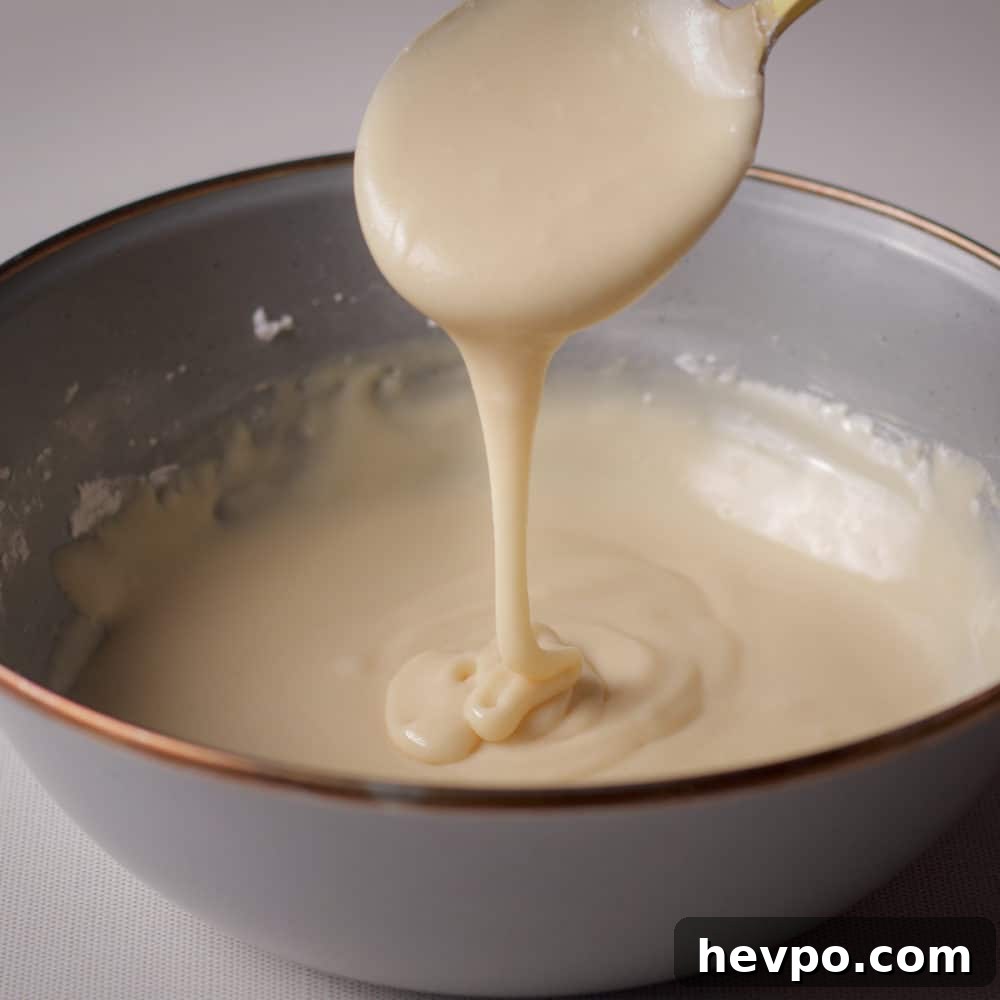 Lifting a spoon from the bowl of icing to show a pourable consistency.
