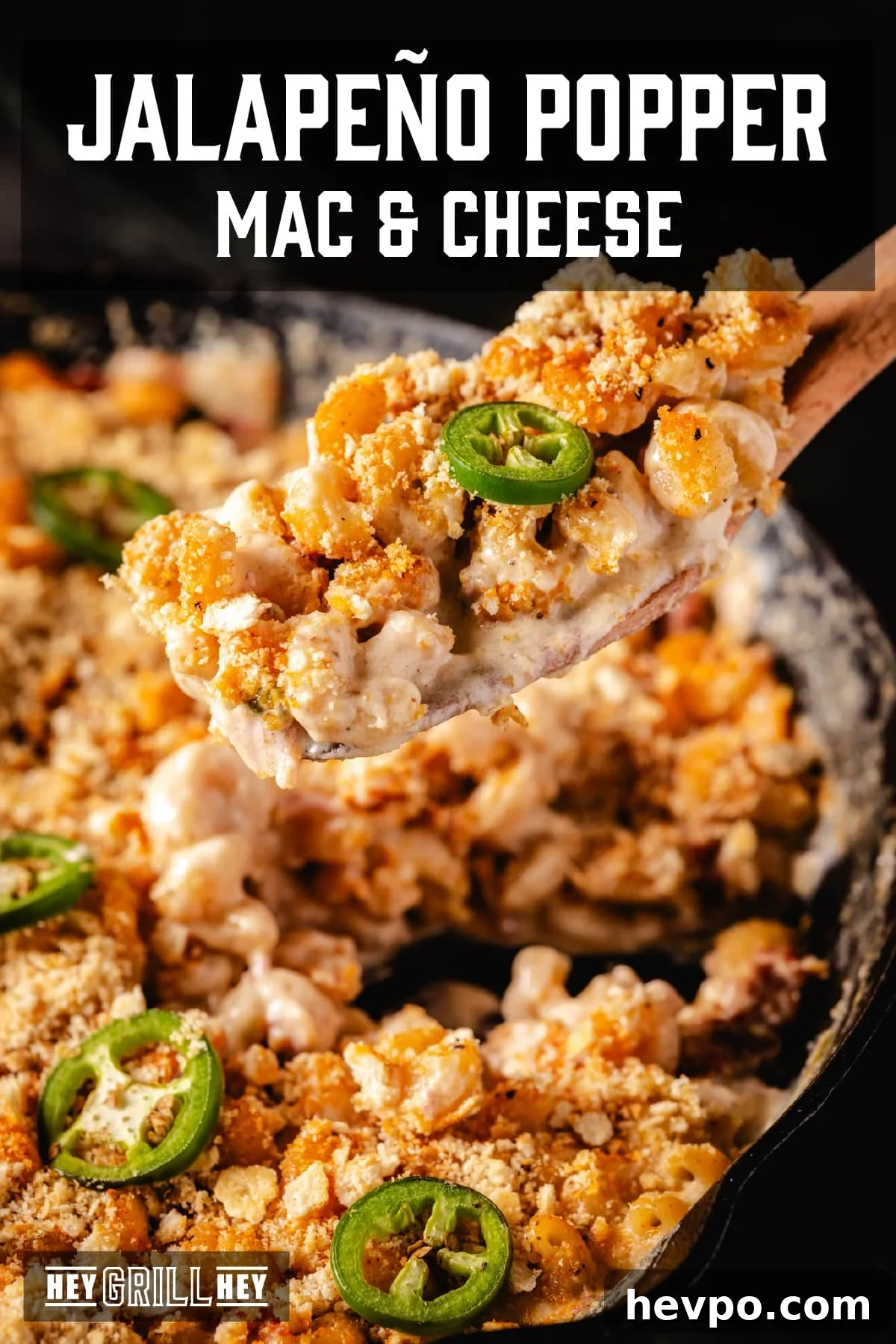 Smoky Jalapeno Pop and Cheese A wooden spatula holding a scoop of jalapeño mac and cheese above a skillet of the same dish. The text overlay reads "Jalapeño Popper Mac & Cheese" at the top, and "Hey Grill Hey" at the bottom.