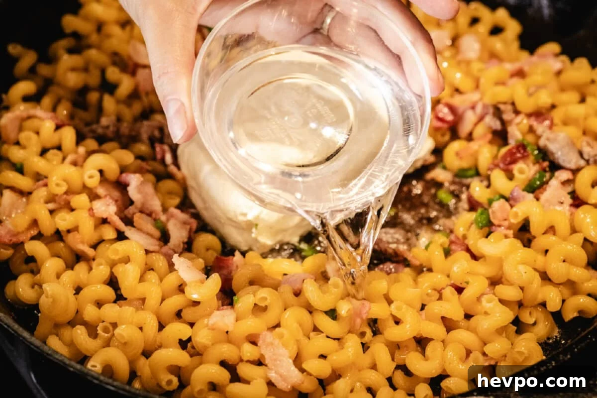Jalapeño Popper Macaroni and Cheese Recipe 2 A chef's hands pouring a clear liquid from a clear bowl into a skillet filled with pasta noodles.