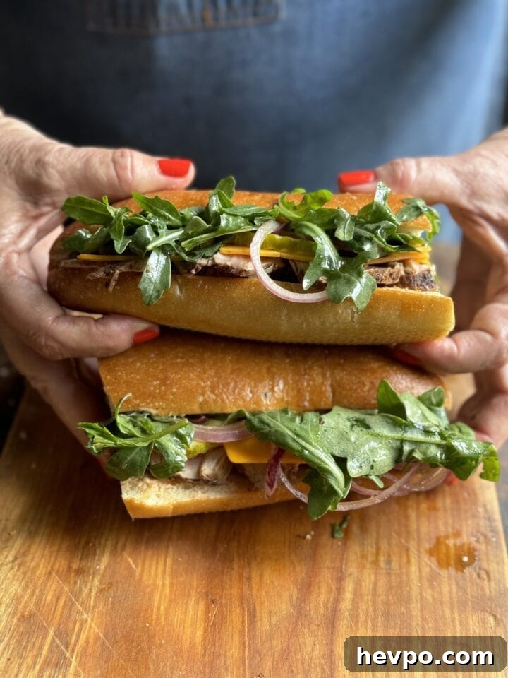 two pork tenderloin sandwiches with arugula cheese and onions on a French roll halves stacked by two hands with red nail polish