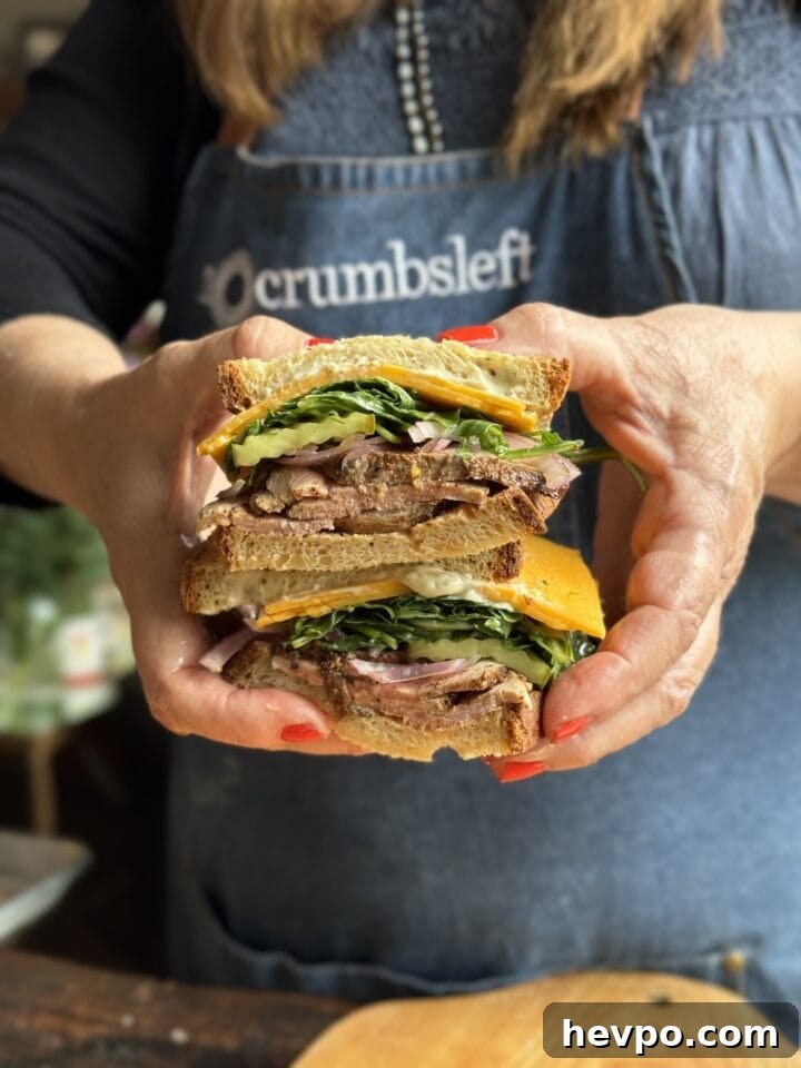 two pork tenderloin sandwich halves with arugula cheese and onions on a sandwich bread stacked by two hands with red nail polish