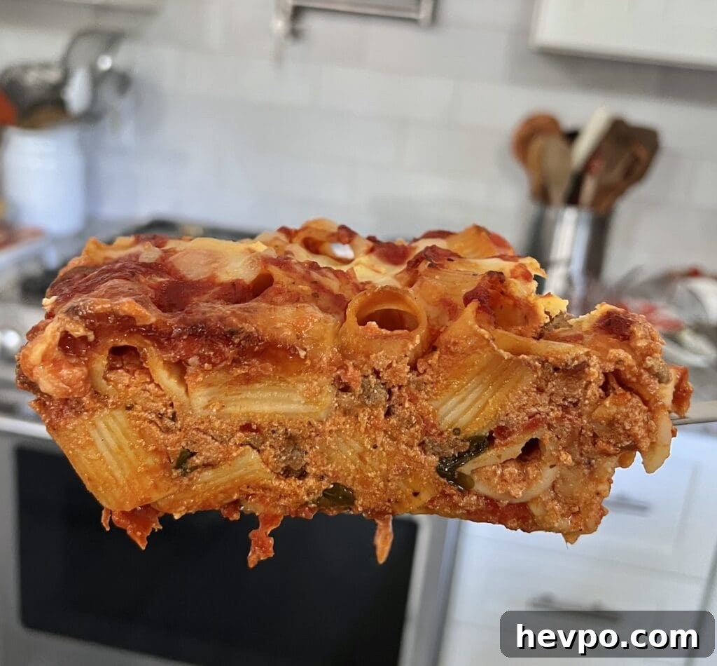 Baked Rigatoni al Forno with Sausage and Beef: Rustic Oven Pasta 3 Side view of a large slice of pasta al forno on a spatula with kitchen in the background.