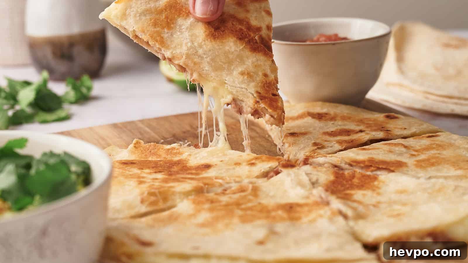 31 Copycat Restaurant Recipes for Delicious Nights In 15 A person lifts a cheesy quesadilla slice from a wooden board. Nearby are bowls of guacamole and salsa on a light-colored table.