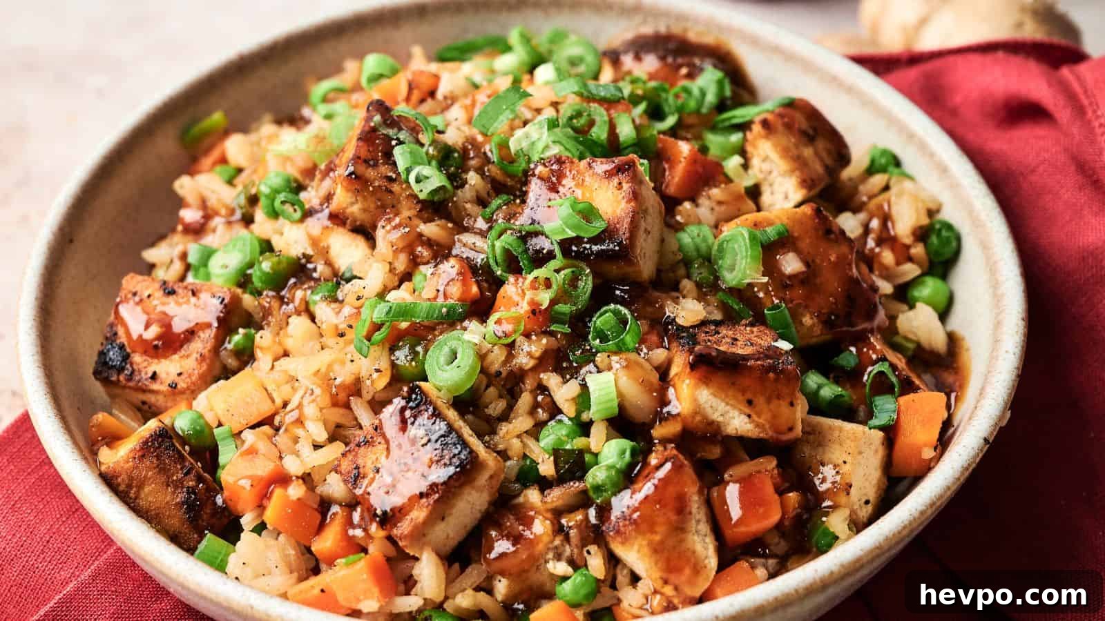 31 Copycat Restaurant Recipes for Delicious Nights In 25 A bowl of fried rice with grilled tofu, diced carrots, peas, chopped green onions, and a drizzle of dark sauce, served on a red cloth.