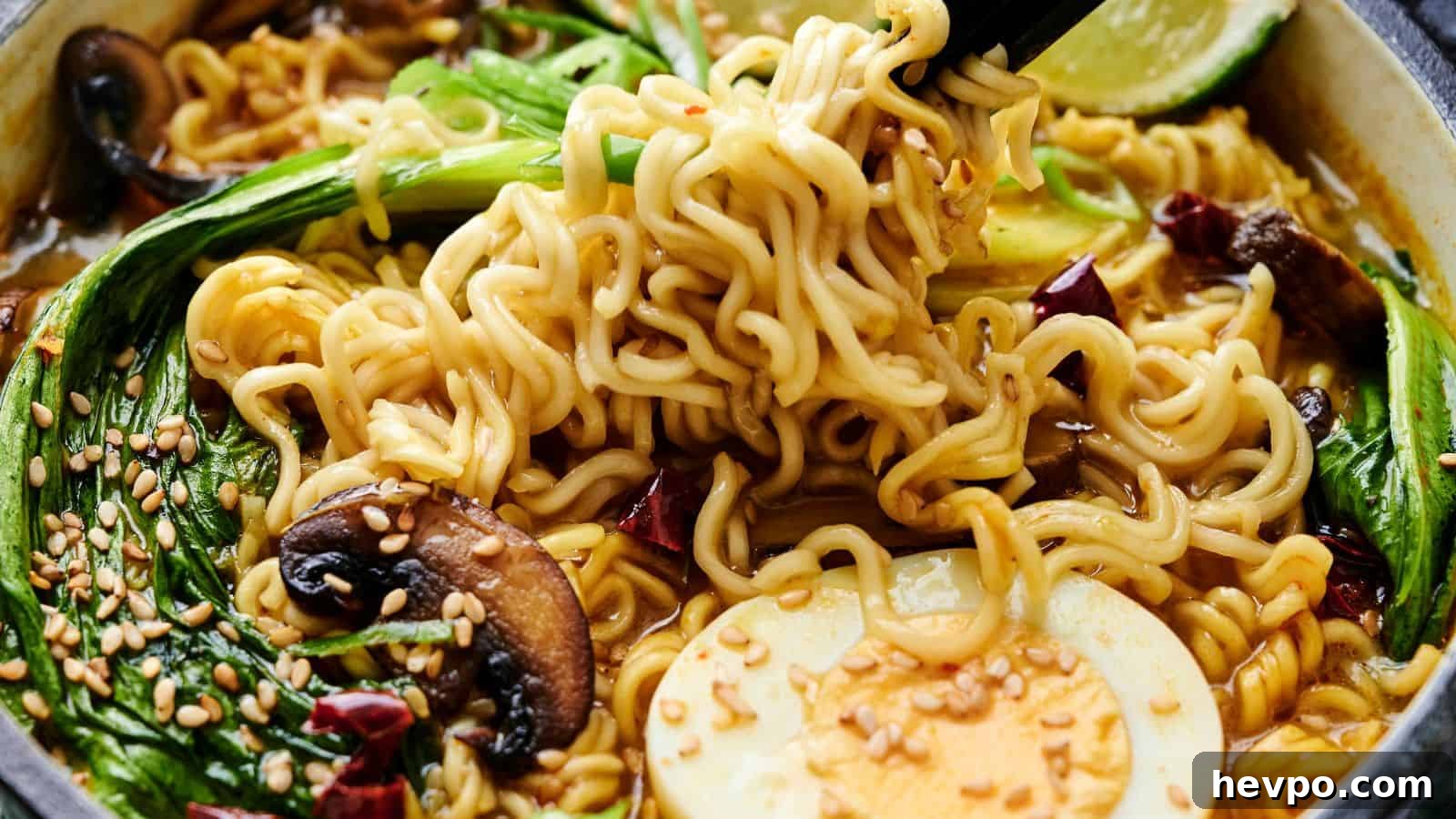 31 Copycat Restaurant Recipes for Delicious Nights In 6 Close-up of a bowl of ramen with noodles, bok choy, mushrooms, half a boiled egg, sesame seeds, and lime, garnished with chili slices. Black chopsticks hold some noodles above the bowl.