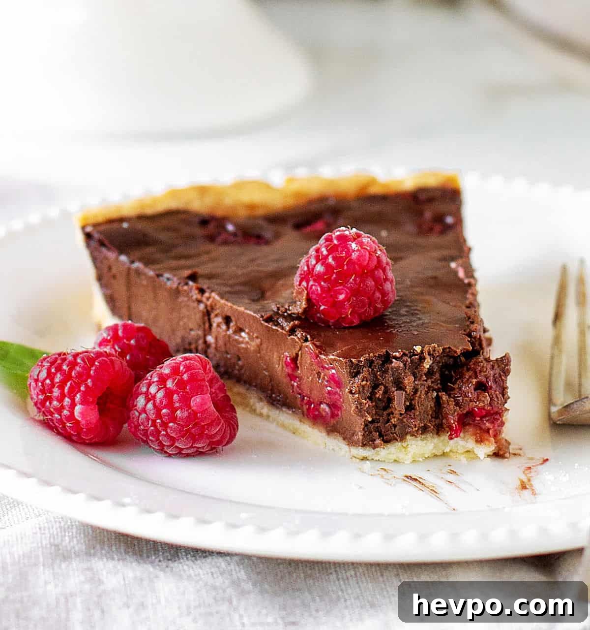 Slice of chocolate raspberry tart with missing bite, on white plate, silver fork.