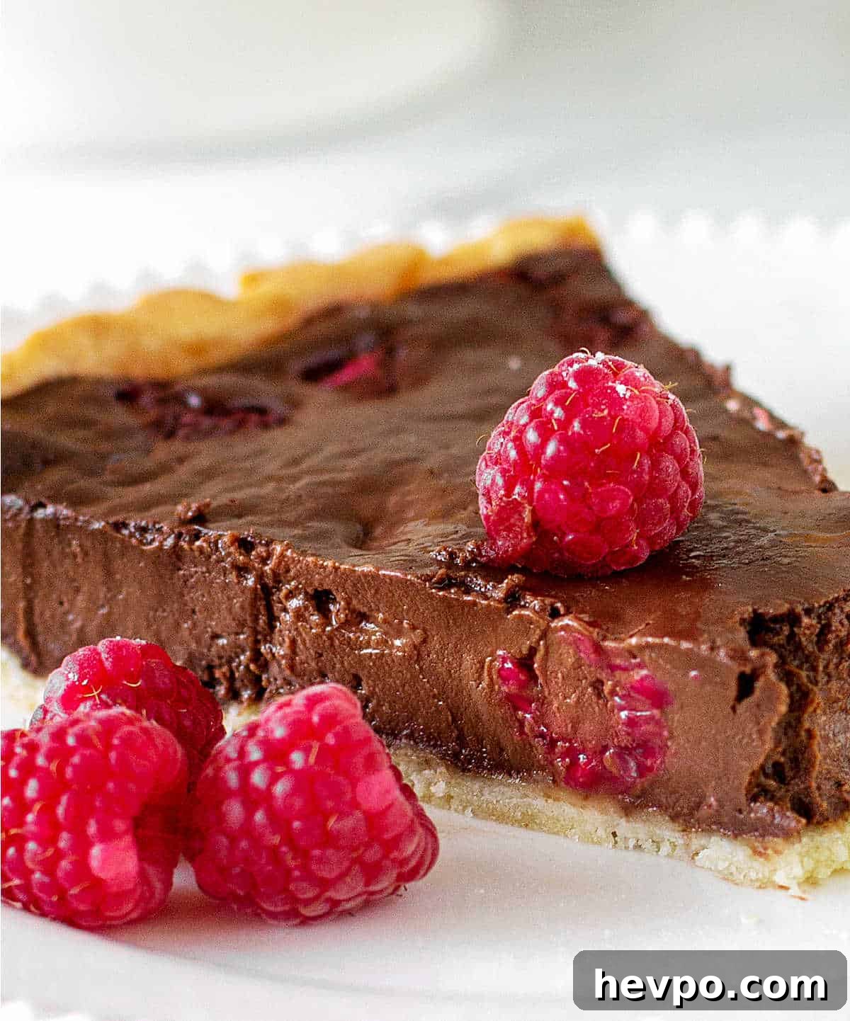 A close up slice of chocolate raspberry tart on a white plate with fresh raspberries.