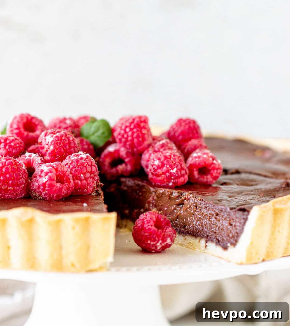 Raspberry chocolate tart on a white cake stand with a missing slice. Fresh raspberries on top. White background.