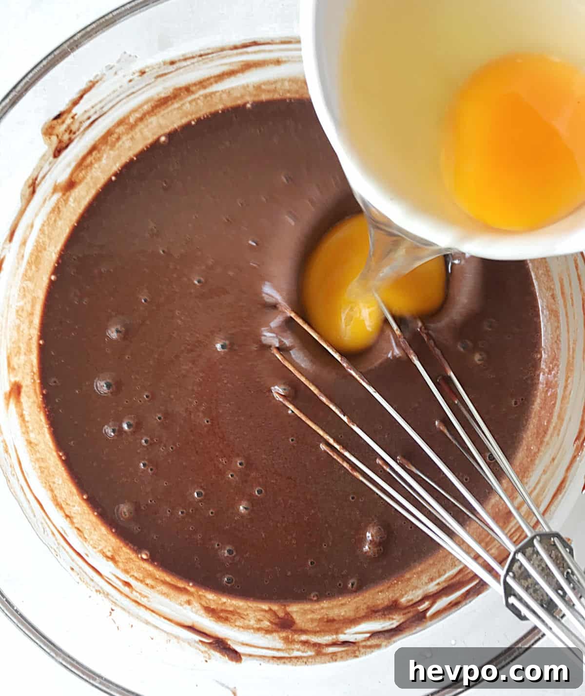 Adding eggs to chocolate mixture in a glass bowl with a whisk inside. White surface, top view.