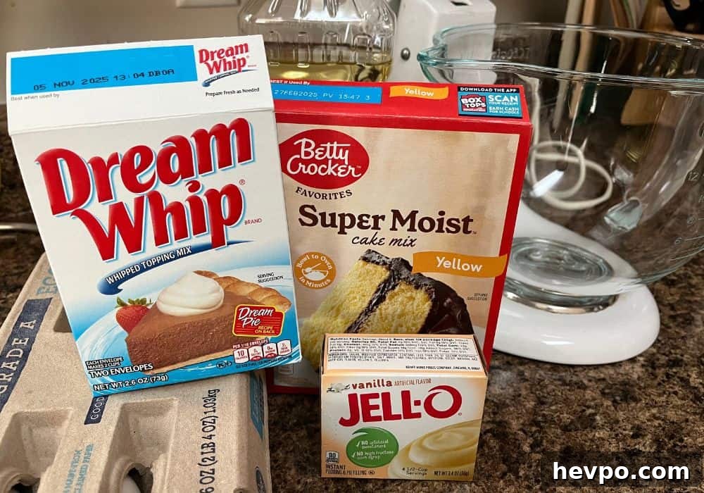 some of the ingredients needed for Dream Whip cake, including Dream Whip mix, cake mix, pudding mix, eggs, and oil. A stand mixer is in the background.