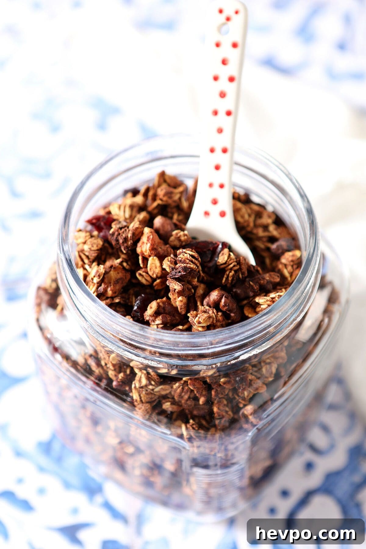 A jar of homemade maple nut granola with a spoon in it on a blue tile surface
