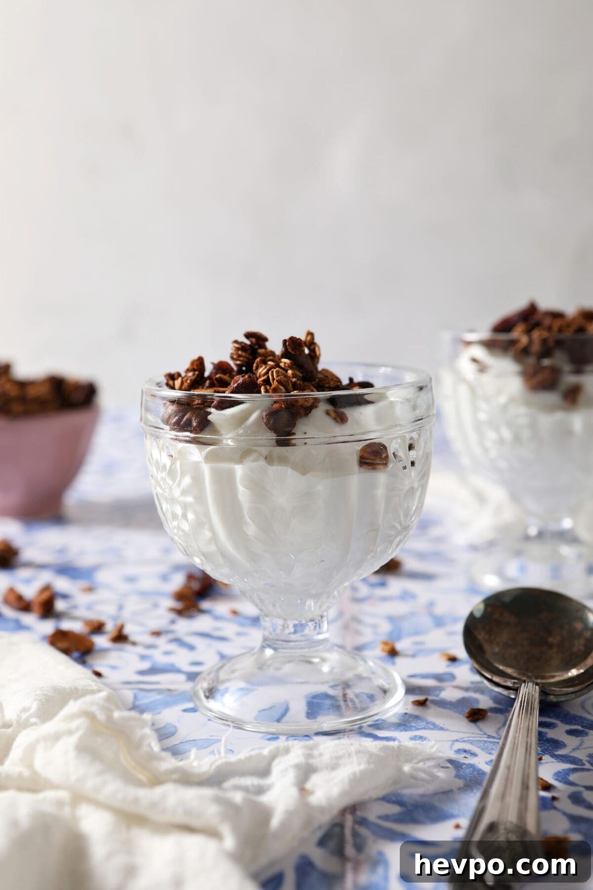 Two glass containers of yogurt topped with granola on a blue tiled surface with spoons