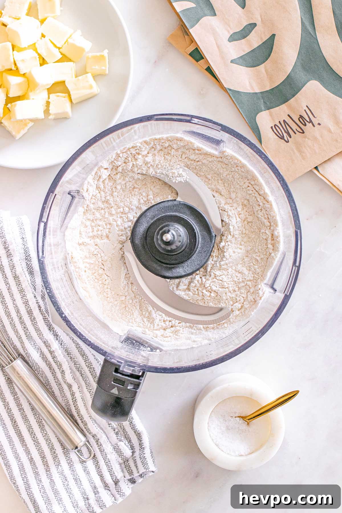 Food processor with flour inside, butter cubes on a plate, and a small bowl of salt on a countertop—everything ready to make Starbucks Copycat Vanilla Bean Scones.