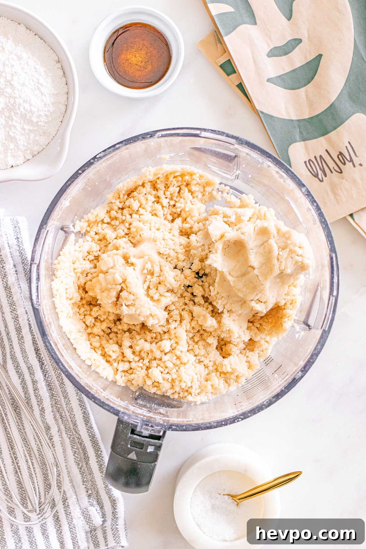 Overhead view of a food processor with cookie dough for Starbucks Copycat Vanilla Bean Scones, sugar, salt, vanilla, and a striped towel on a white surface.