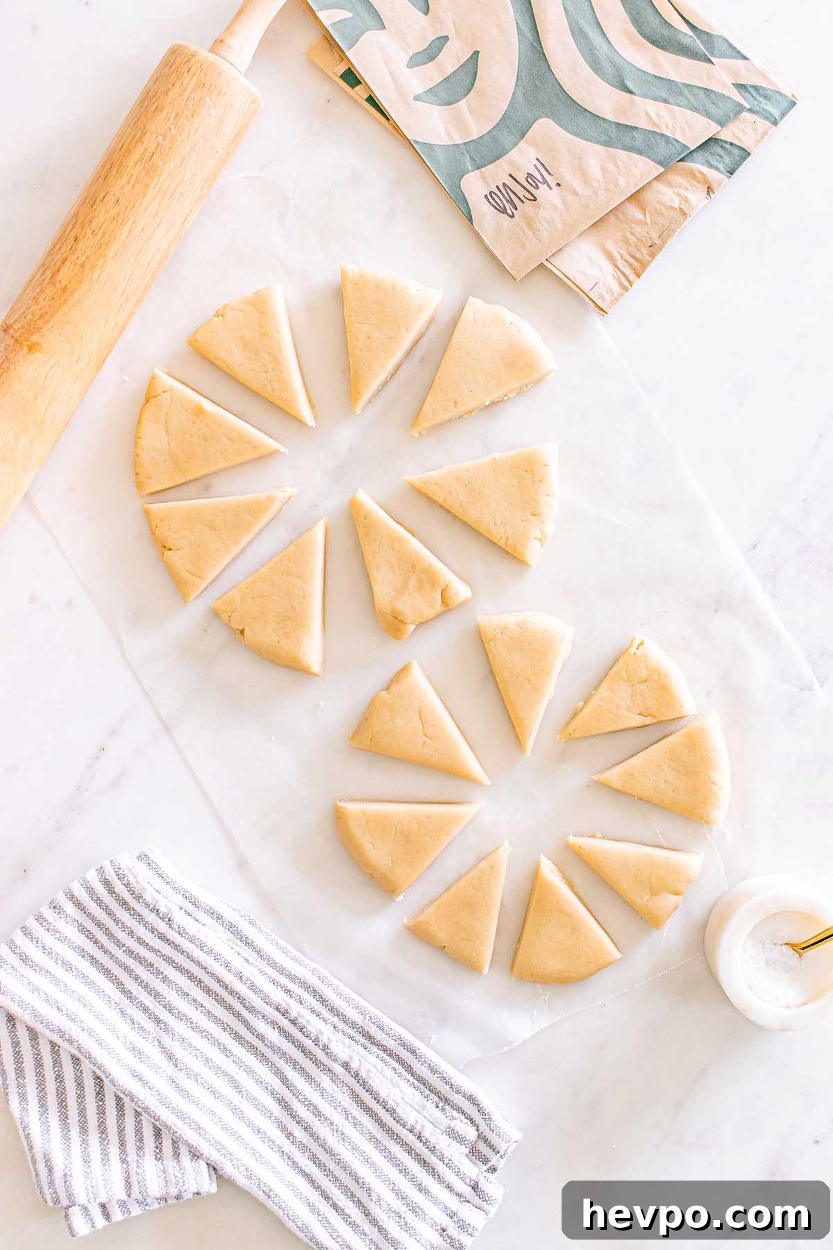 Two circles of dough cut into wedges on parchment, ready to become Starbucks Copycat Vanilla Bean Scones, with a rolling pin, striped towel, and paper bag nearby.