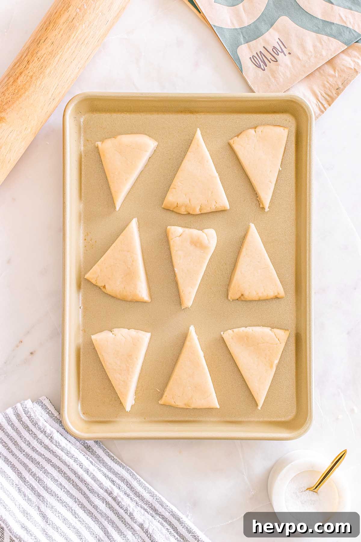 Nine unbaked triangular Starbucks Copycat Vanilla Bean Scones on a baking sheet, ready to bake, with baking tools nearby.