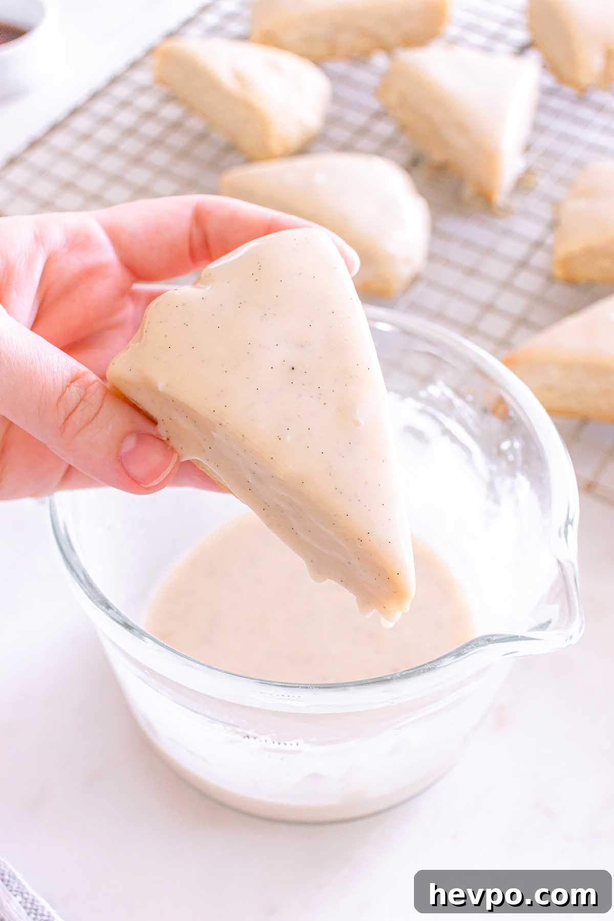 Hand dipping a triangle Starbucks Copycat Vanilla Bean Scone into white glaze, with more scones cooling on a rack in the background.