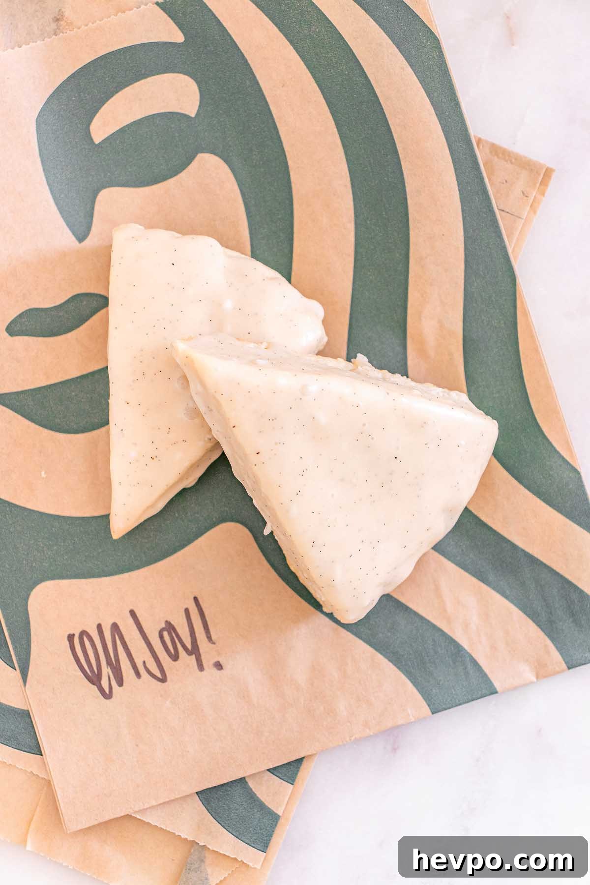 Two Starbucks Copycat Vanilla Bean Scones with icing rest on Starbucks-branded brown paper bags, one featuring the word "enjoy!".