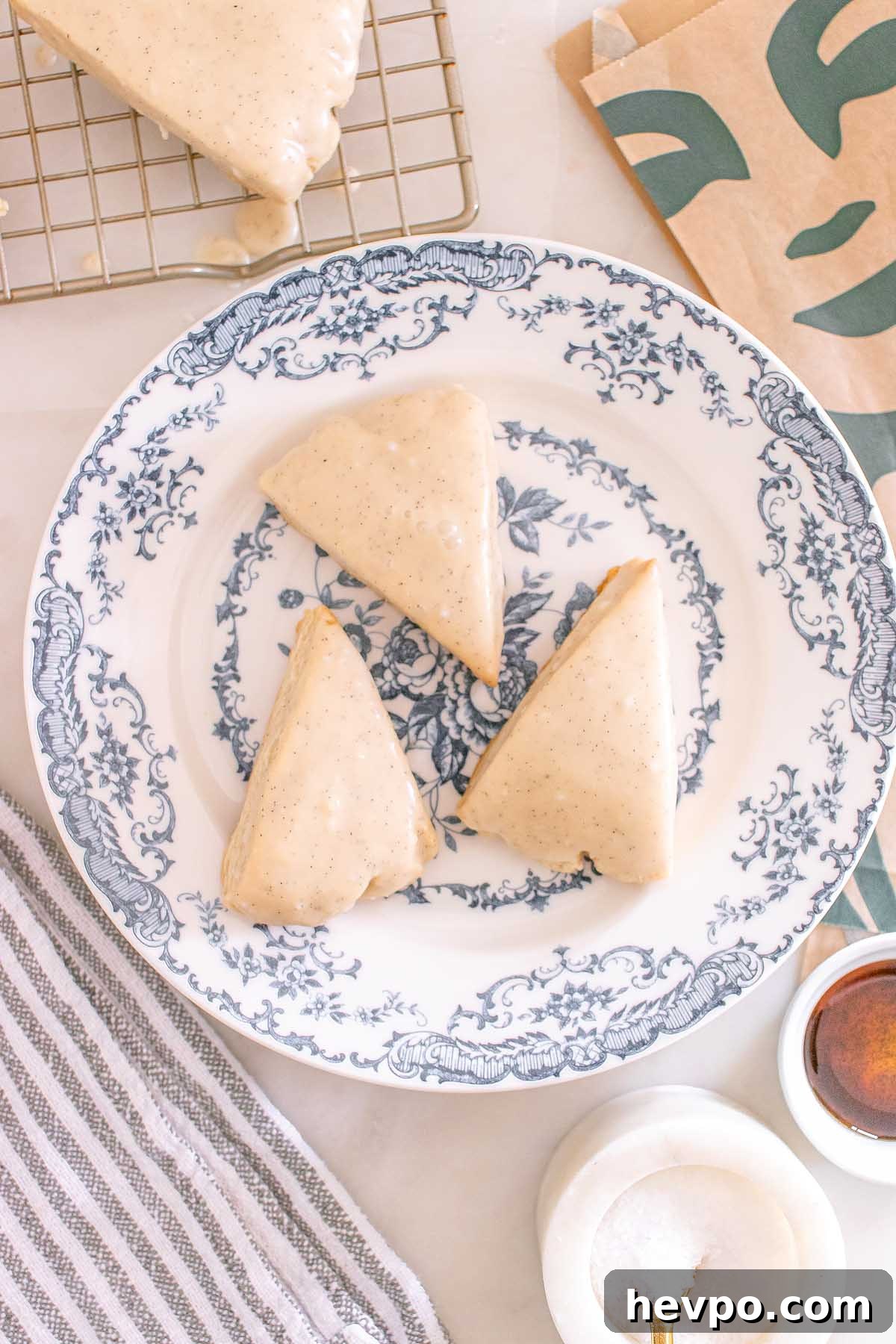 Three glazed Starbucks Copycat Vanilla Bean Scones rest on a blue and white floral plate, with baking items and a striped towel nearby.