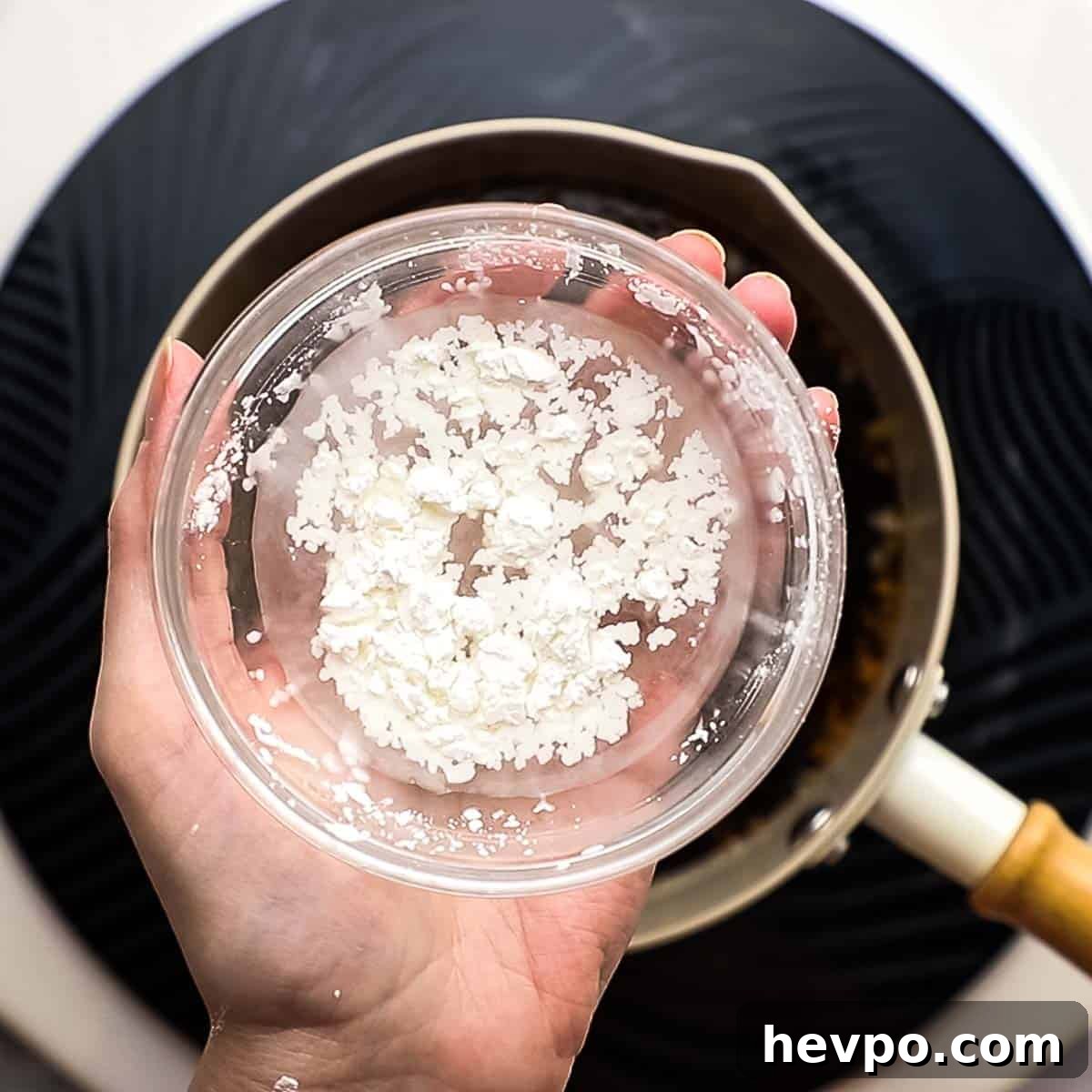 a hand holding a bowl of cornstarch and water over a saucepan
