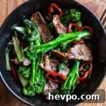wooden background with black bowl of stir fried broccolini, strips of juicy seared sirloin steak and slices of red chilli