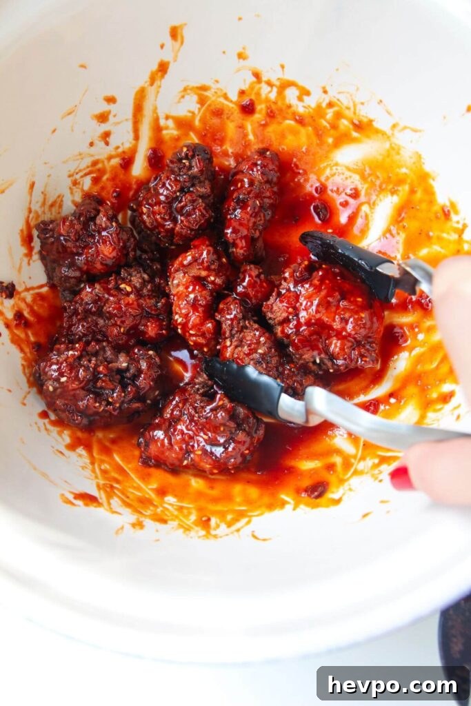 Crispy Korean Fried Chicken Recipe: Extra Crunch & Sticky Sauce 11 Tongs mixing fried chicken with sticky glaze in a large white bowl.