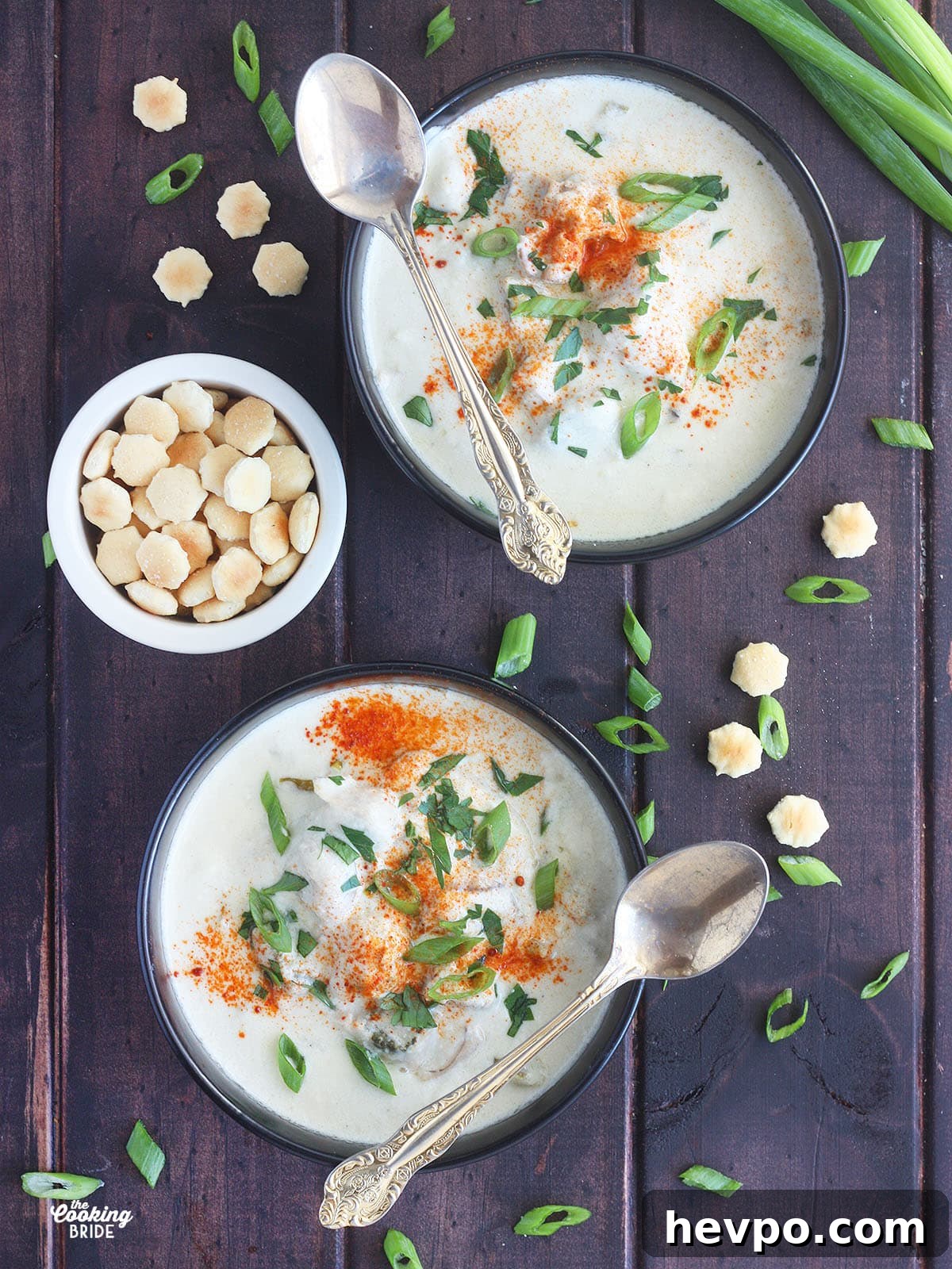 overhead shot of two bowls of oyster stew garnished with green onions and cayenne pepper served in black bowls with spoons an a bowl of oyster crackers resting on the side
