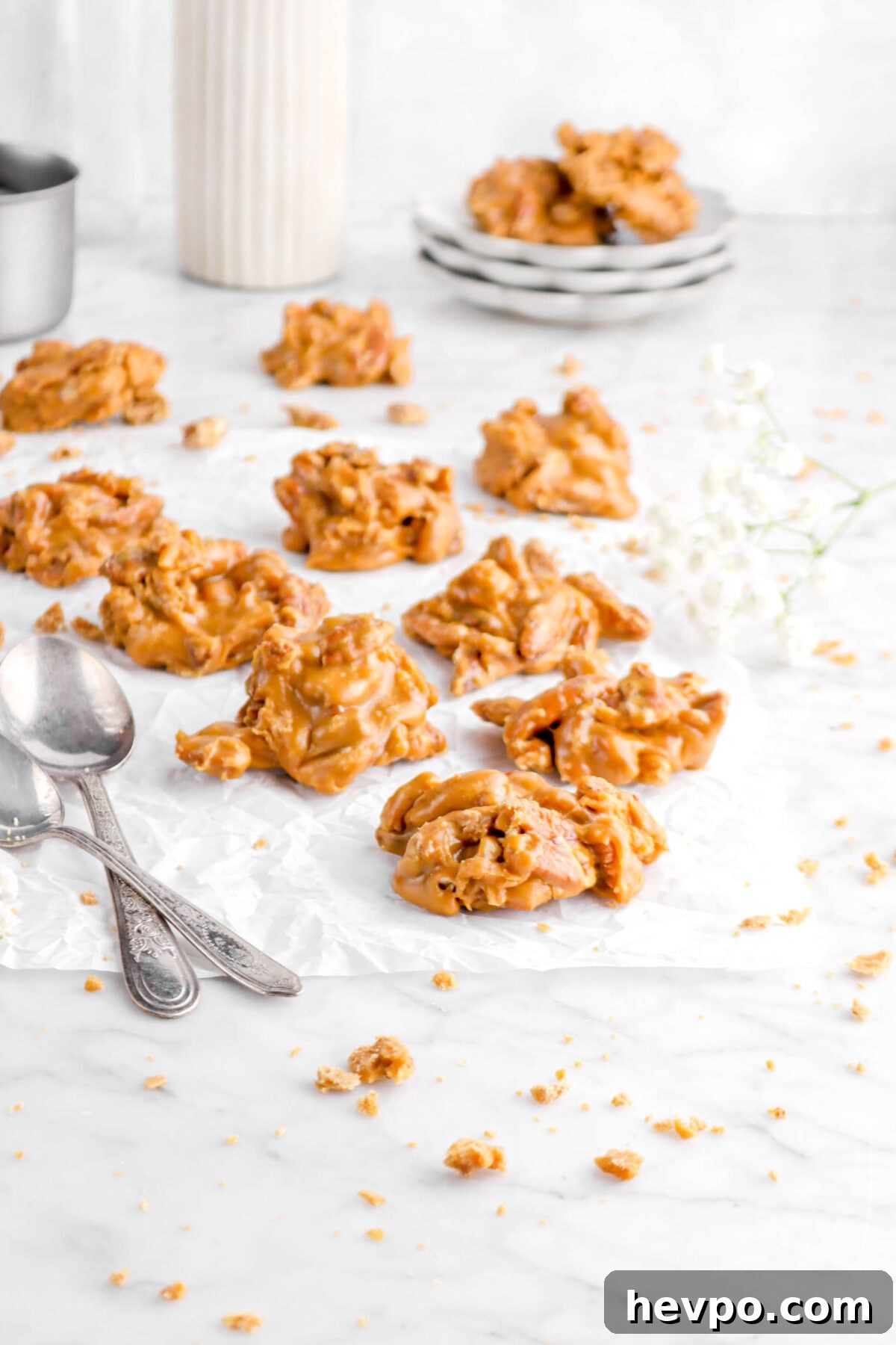 Southern Buttermilk Pecan Pralines Recipe – Classic Southern Candy 15 pulled back shot of pecan pralines on parchment with pralines crumbles around, two spoons, flowers, and stack of plates behind
